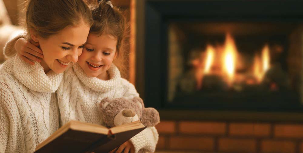 Mother and Child In Sweaters reading a book with a fireplace in the Background